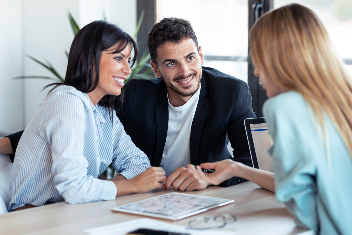 Shot of beautiful real-estate agent showing house plans on electronic tablet while talking to the couple about buying the house in the office